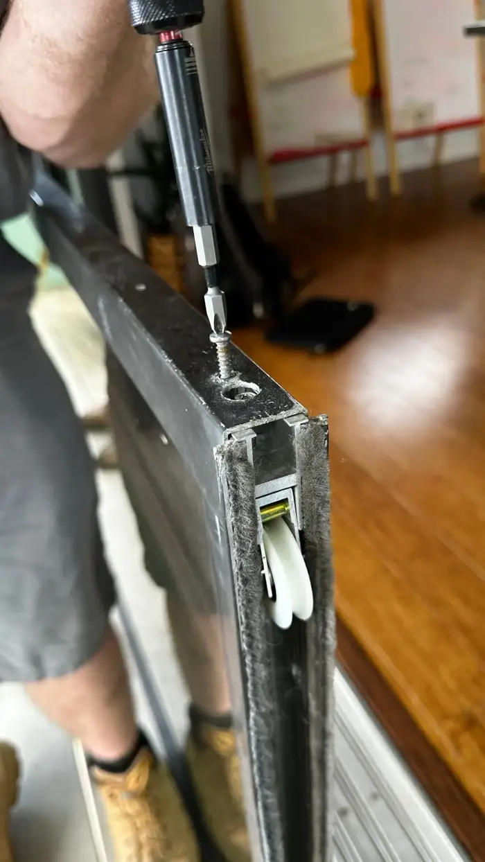 Technician using a Milwaukee impact driver to remove the bottom roller screw from a sliding glass door panel
